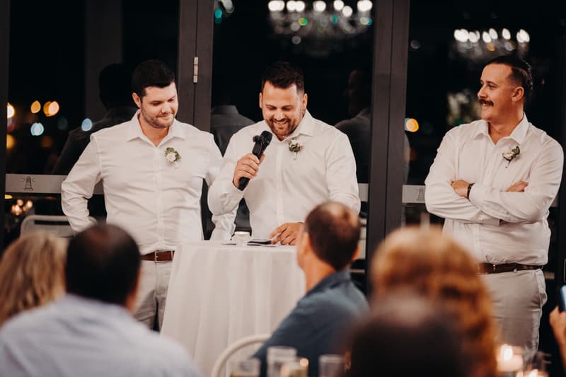Brodie, the groom, stands at a high table holding a microphone and speaking during the reception at Sandstone Point Hotel — Pumicestone Room, flanked by two groomsmen in white shirts with boutonnieres, while guests listen in the foreground.
