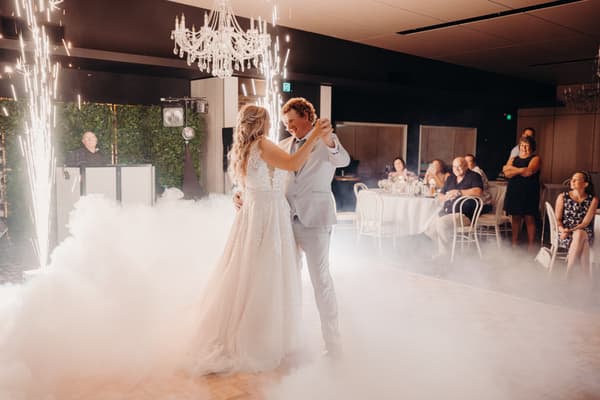 The bride Chloe and groom Brodie share their first dance surrounded by low fog and sparklers at the Sandstone Point Hotel — Pumicestone Room, with seated and standing guests watching in the background.