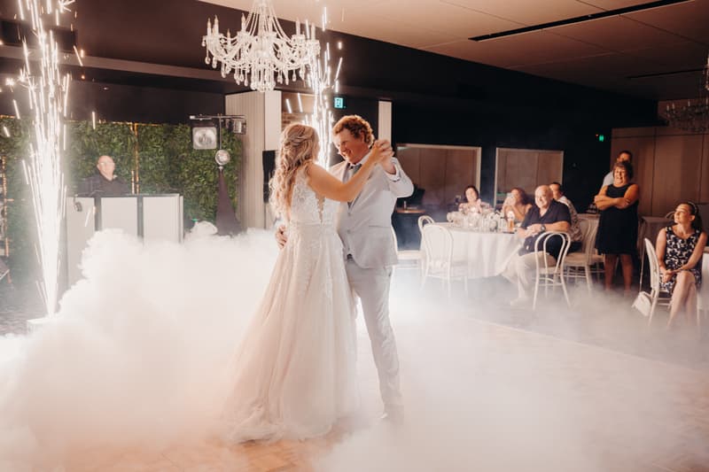 The bride Chloe and groom Brodie share their first dance surrounded by low fog and sparklers at the Sandstone Point Hotel — Pumicestone Room, with seated and standing guests watching in the background.