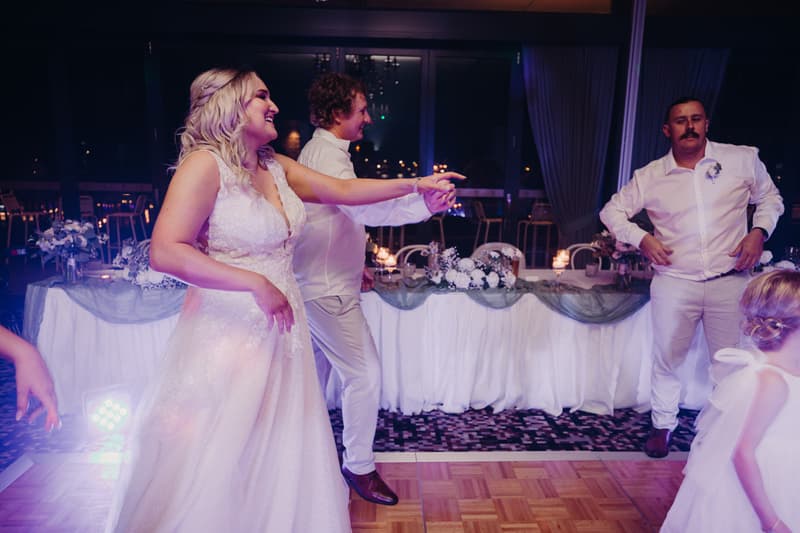 The bride Chloe and the groom Brodie dance near the head table decorated with floral arrangements and candles at Sandstone Point Hotel — Pumicestone Room during the wedding reception.