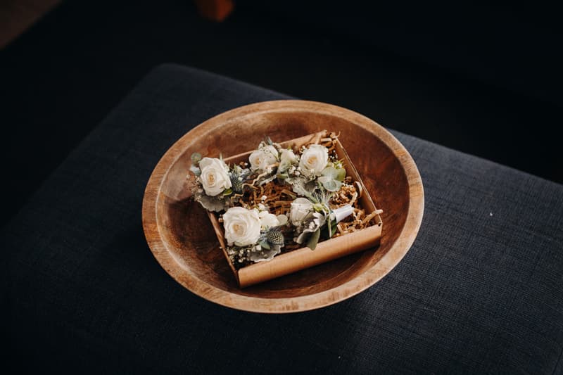A wooden bowl containing a small box with four white rose boutonnieres resting on shredded paper filler.