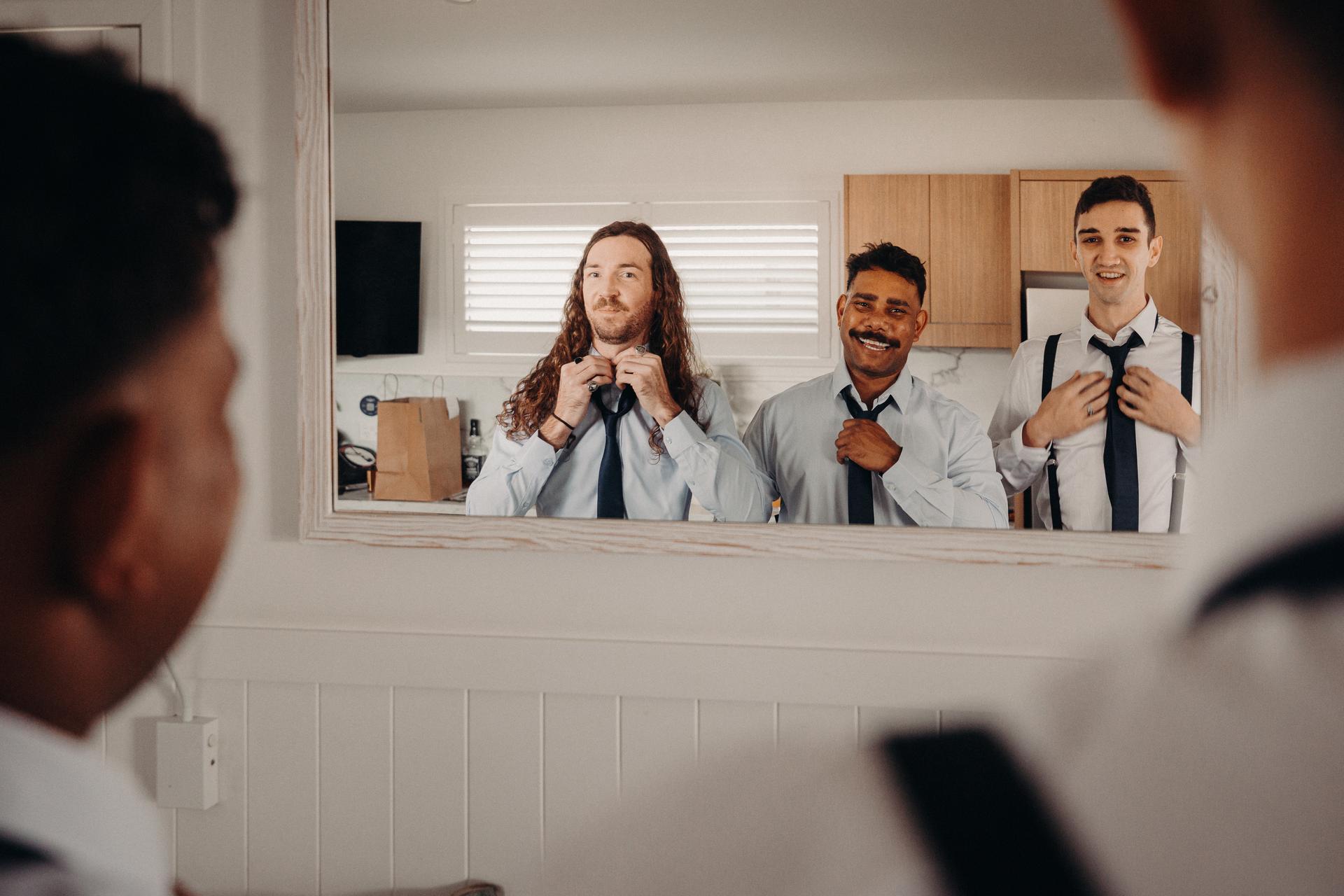 Three groomsmen adjust their ties while looking into a mirror in a room at Sandstone Point Hotel.