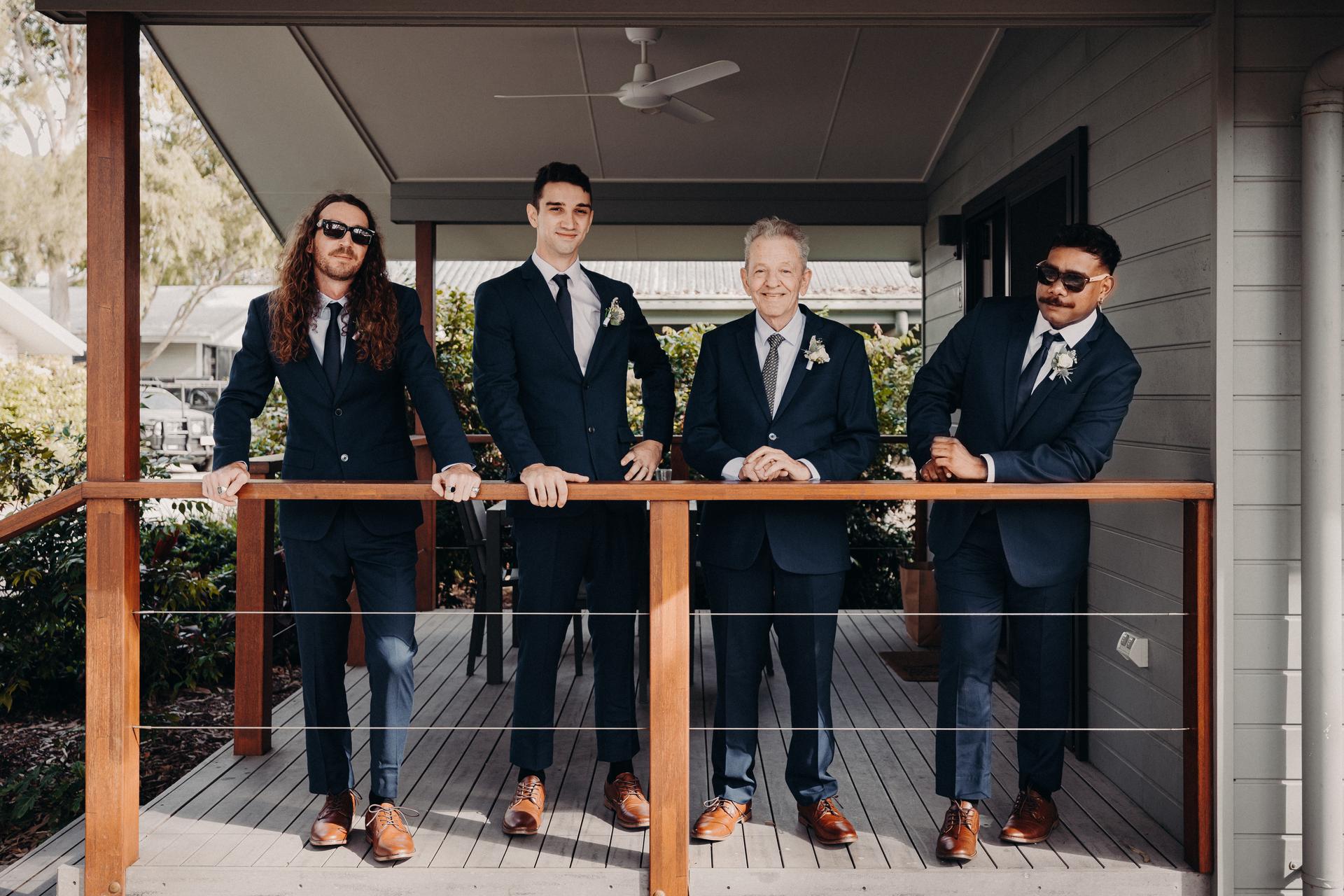 Four men in navy suits with boutonnieres stand on the porch of Sandstone Point Hotel Pavilion, leaning on the wooden railing.
