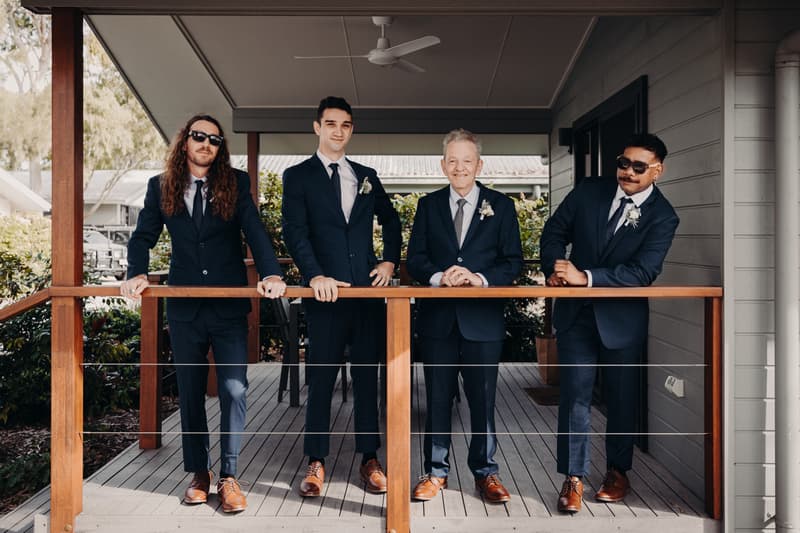 Four men in navy suits with boutonnieres stand on the porch of Sandstone Point Hotel Pavilion, leaning on the wooden railing.