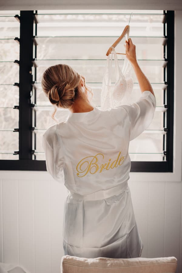 The bride stands with her back to the camera holding a lace wedding dress on a hanger near a window with black slatted blinds.