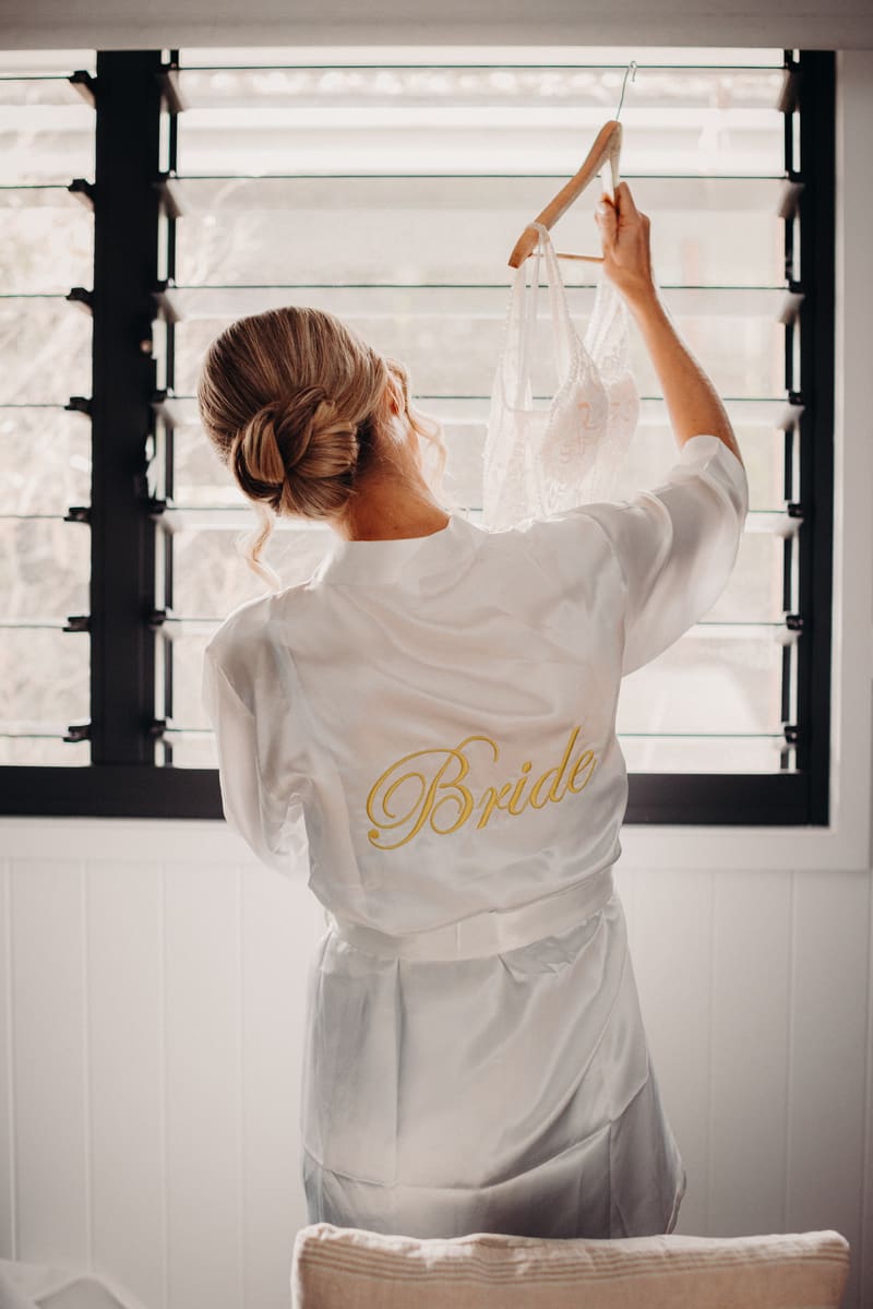 The bride stands with her back to the camera holding a lace wedding dress on a hanger near a window with black slatted blinds. Gold Coast wedding videographer by She Said Yes.