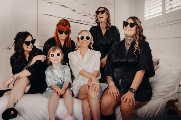 The bride Courtney in a white robe and four bridesmaids in black robes, along with a flower girl in a light blue robe, all wearing sunglasses, sit together on a bed in a room at Sandstone Point Hotel — Pavilion.