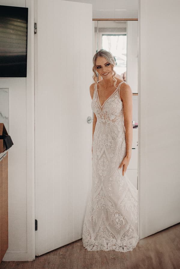 The bride Courtney stands in a doorway wearing a detailed lace wedding gown inside a preparation room at Sandstone Point Hotel.