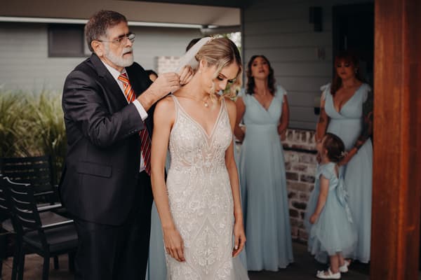The bride Courtney stands as an older man, likely her father, adjusts her veil. Three bridesmaids and a flower girl in light blue dresses stand nearby inside the Sandstone Point Hotel Pavilion.