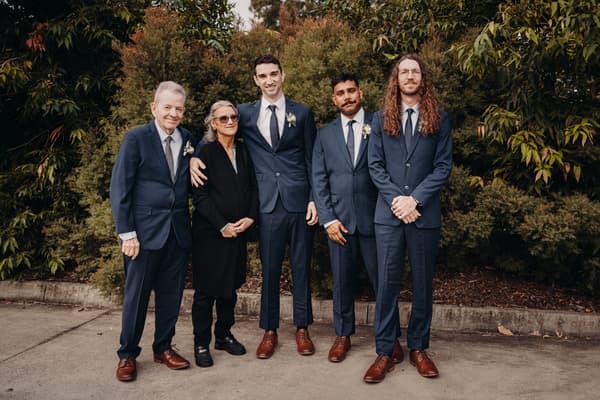 Groom Cameron stands with four guests, including an older man and woman, outside at Sandstone Point Hotel — Pavilion, all dressed formally with suits and boutonnieres.