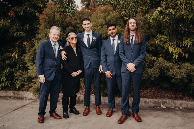Groom Cameron stands with four guests, including an older man and woman, outside at Sandstone Point Hotel — Pavilion, all dressed formally with suits and boutonnieres.