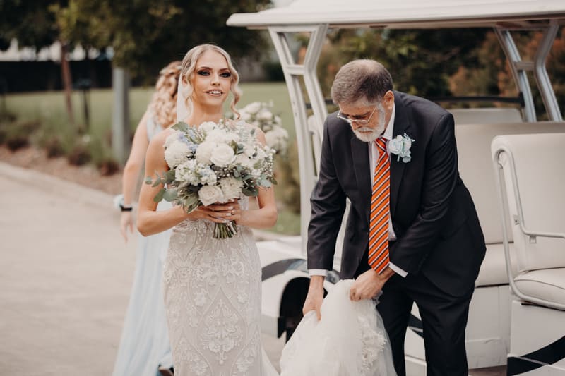 The bride Courtney holds a bouquet of white and light blue flowers while an older man, likely the bride's father, adjusts her wedding dress near a white golf cart. Photo and video packages by She Said Yes.
