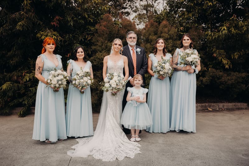 Courtney the bride stands with bridesmaids in light blue dresses, a flower girl, and an older man. Photo and video packages by She Said Yes.