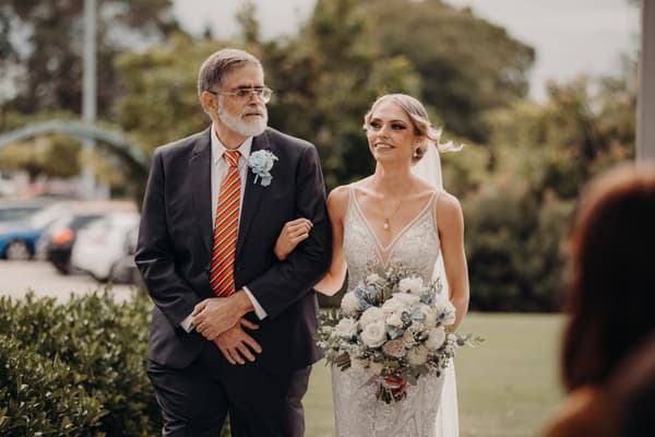 The bride Courtney is walking arm-in-arm with an older man, likely her father, outside the Sandstone Point Hotel — Pavilion, holding a bouquet of white and blue flowers.