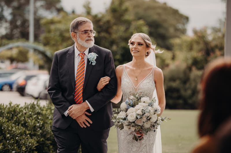The bride Courtney is walking arm-in-arm with an older man, likely her father, outside the Sandstone Point Hotel — Pavilion, holding a bouquet of white and blue flowers.