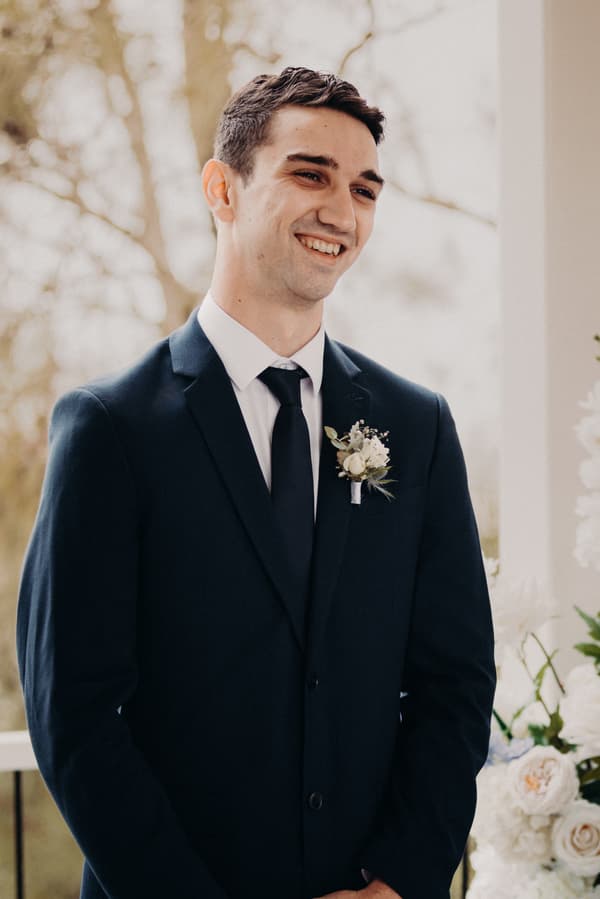 The groom Cameron stands smiling at the Sandstone Point Hotel — Pavilion during the ceremony stage, wearing a dark suit with a boutonniere.