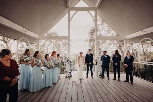 Courtney the bride and Cameron the groom stand at the ceremony stage at Sandstone Point Hotel — Pavilion, flanked by bridesmaids in light blue dresses holding bouquets on the left and groomsmen in dark suits on the right, with floral arrangements and trees visible in the background.