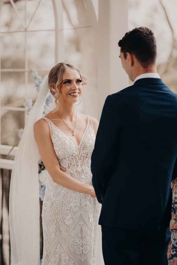 Courtney the bride and Cameron the groom stand facing each other at the ceremony stage inside the Sandstone Point Hotel — Pavilion, with Courtney wearing a detailed lace wedding dress and veil, and Cameron in a dark suit.