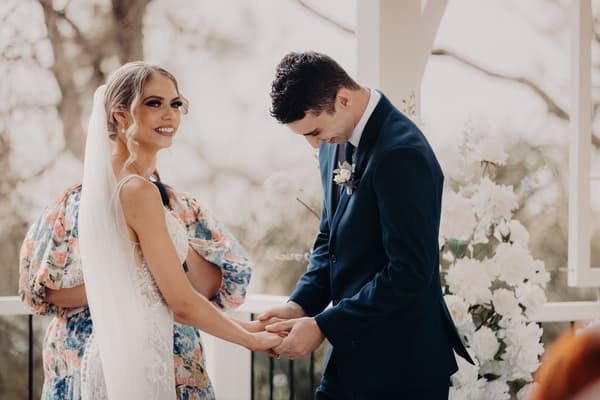 Courtney the bride and Cameron the groom hold hands during their wedding ceremony at Sandstone Point Hotel — Pavilion, with a floral arrangement visible in the background.