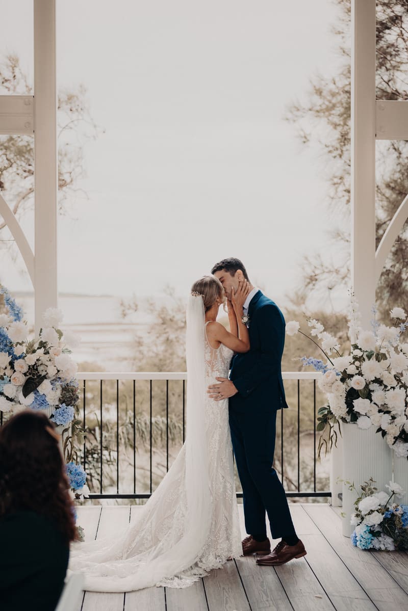 Bride Courtney and groom Cameron share a kiss at the ceremony stage of, surrounded by floral arrangements and an outdoor backdrop. Brisbane wedding video by She Said Yes.