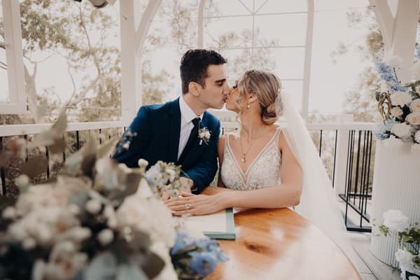 Courtney and Cameron kiss while seated at a wooden table during their ceremony at Sandstone Point Hotel — Pavilion, with floral arrangements in the foreground and background.
