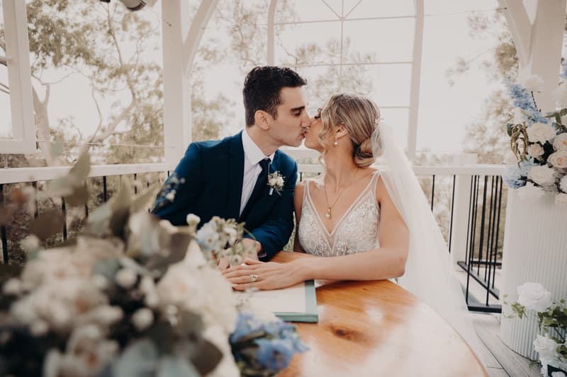 Courtney and Cameron kiss while seated at a wooden table during their ceremony at Sandstone Point Hotel — Pavilion, with floral arrangements in the foreground and background.