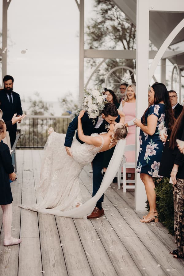 Courtney and Cameron share a kiss on the ceremony stage at Sandstone Point Hotel — Pavilion as guests watch and celebrate.