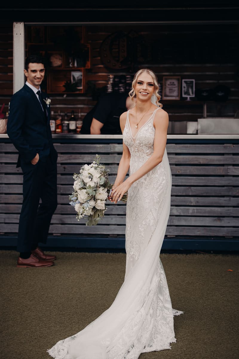 The bride Courtney stands holding a bouquet of white flowers in front of a wooden bar area, while the groom Cameron stands in the background wearing a dark suit. Full day wedding photography by She Said Yes.