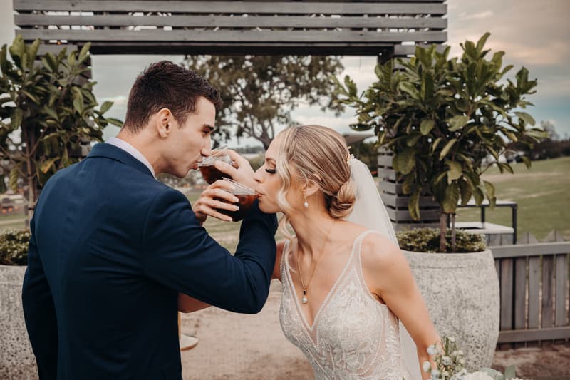 Courtney the bride and Cameron the groom drink from plastic cups while linking arms at Sandstone Point Hotel during their couple portraits session.