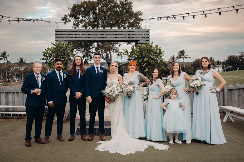 Courtney the bride and Cameron the groom pose with their wedding party at Sandstone Point Hotel, including bridesmaids in light blue dresses, groomsmen in navy suits, and a flower girl in a light blue dress, standing outdoors under string lights.