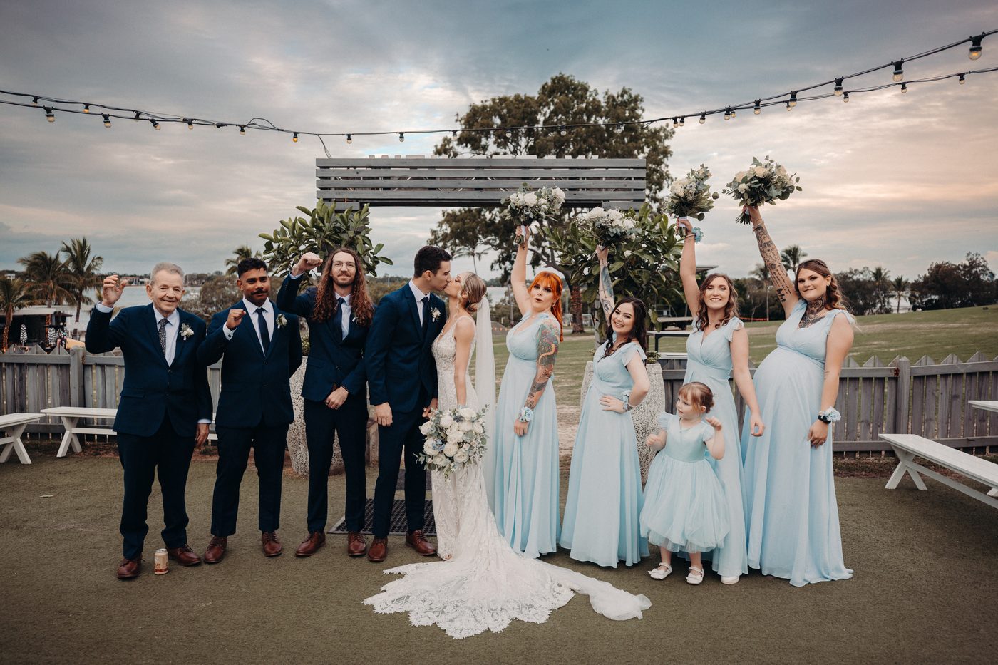 Courtney and Cameron kiss during couple portraits, surrounded by bridesmaids in light blue dresses and groomsmen in navy suits, with a flower girl and an older man also present. How much does a wedding photographer cost in Brisbane by She Said Yes.