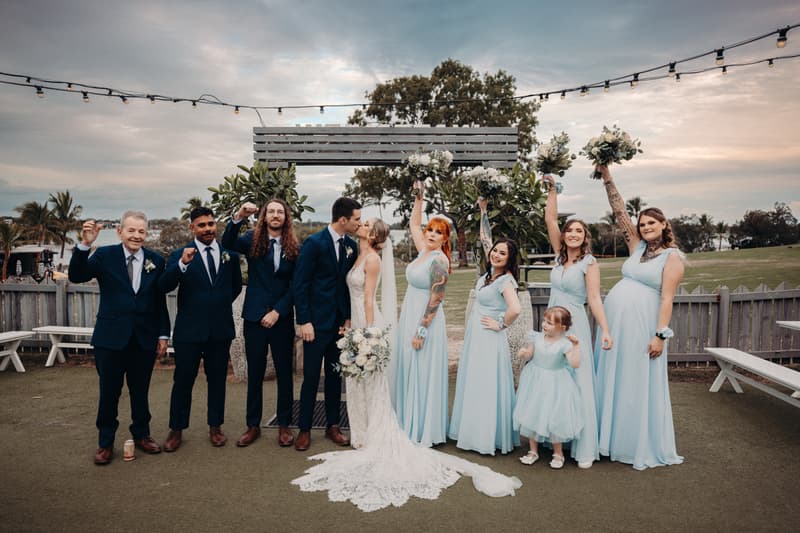 Courtney and Cameron kiss during couple portraits at Sandstone Point Hotel, surrounded by bridesmaids in light blue dresses and groomsmen in navy suits, with a flower girl and an older man also present.