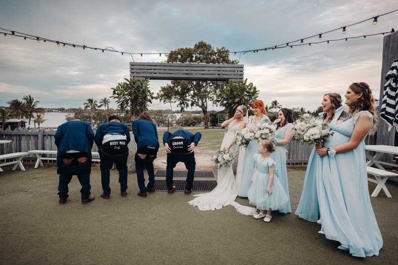 Courtney the bride and bridesmaids in light blue dresses stand holding bouquets and laughing as Cameron the groom and groomsmen bend over showing the backs of their jackets with text at Sandstone Point Hotel outdoor lawn area near water.