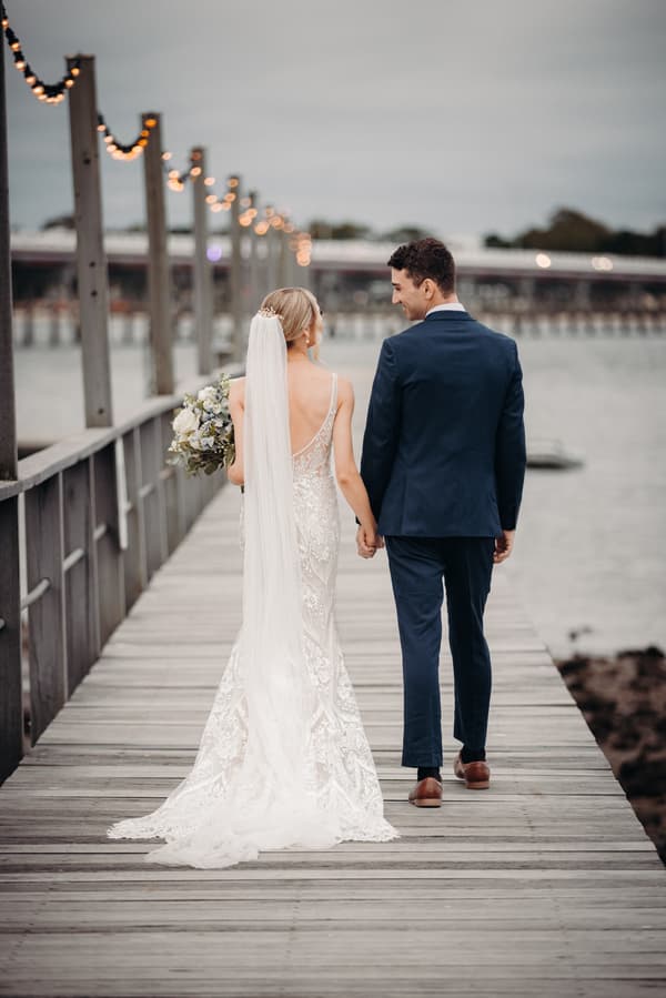 The bride Courtney in a lace wedding gown with a long veil and the groom Cameron in a navy suit walk hand in hand along a wooden pier at Sandstone Point Hotel, with string lights hanging on posts along the pier and water visible on the right.