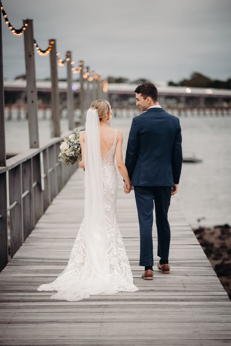 The bride Courtney in a lace wedding gown with a long veil and the groom Cameron in a navy suit walk hand in hand along a wooden pier, with string lights hanging on posts along the pier and water visible on the right. Wedding industry awards finalist by She Said Yes.