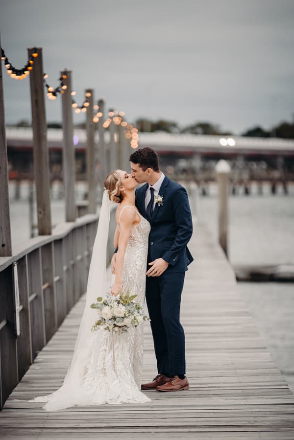 Courtney the bride and Cameron the groom share a kiss on a wooden pier at Sandstone Point Hotel, with the bride holding a bouquet and string lights visible on posts along the pier.