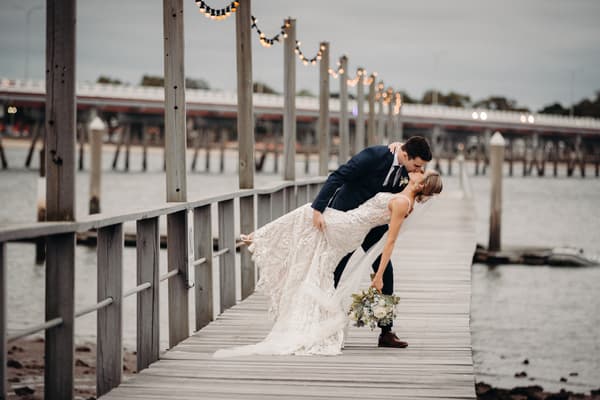 Courtney and Cameron pose for a couple portrait on a wooden jetty at Sandstone Point Hotel, with Cameron dipping Courtney as they kiss. Courtney holds a bouquet of flowers and string lights hang along the jetty posts.