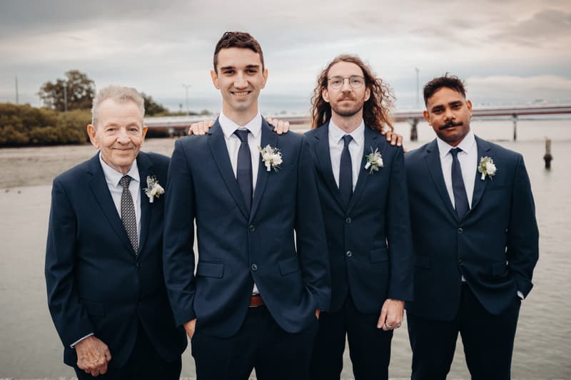 Four men in navy suits with boutonnieres pose together outdoors near water at Sandstone Point Hotel.