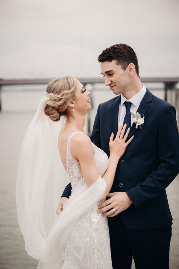 Courtney the bride and Cameron the groom pose together near water at Sandstone Point Hotel during their couple portraits session. Courtney wears a detailed white wedding gown with a veil, and Cameron wears a dark suit with a boutonniere.
