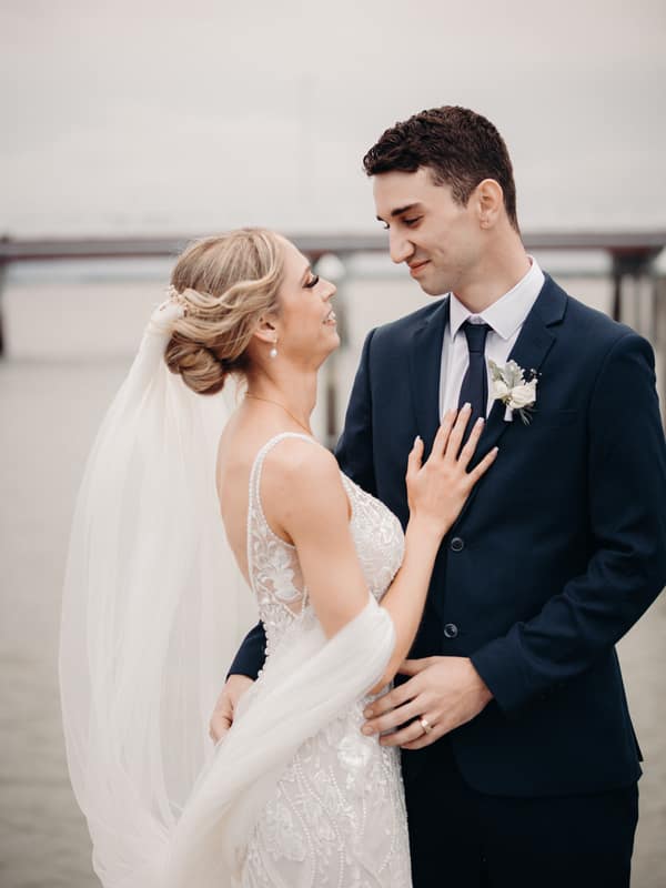 Courtney the bride and Cameron the groom pose together near water at Sandstone Point Hotel during their couple portraits session. Courtney wears a detailed white wedding gown with a veil, and Cameron wears a dark suit with a boutonniere.