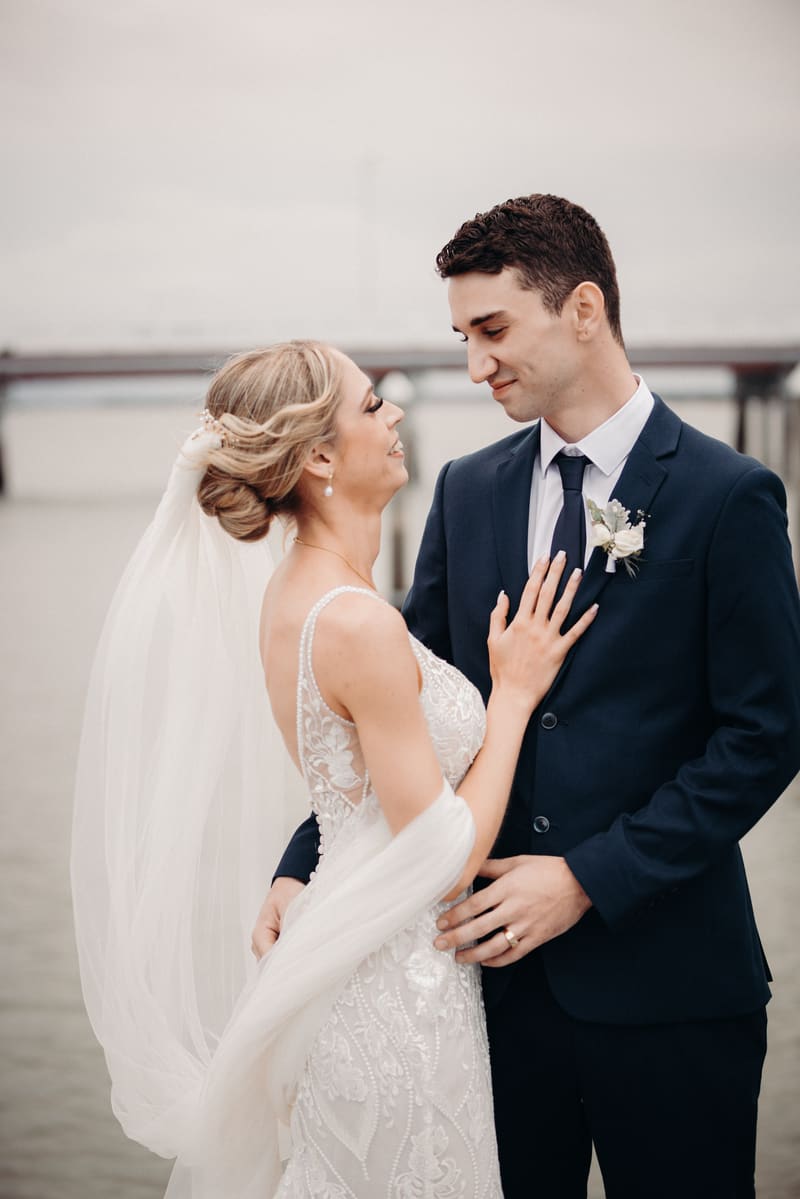 Courtney the bride and Cameron the groom pose together near water. Courtney wears a detailed white wedding gown with a veil, and Cameron wears a dark suit with a boutonniere. Engagement reception by She Said Yes.