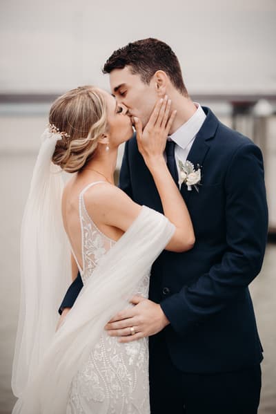 Courtney the bride and Cameron the groom share a kiss during their couple portraits at Sandstone Point Hotel. Courtney wears a white lace wedding gown with a veil and hairpiece, while Cameron wears a dark suit with a boutonniere.