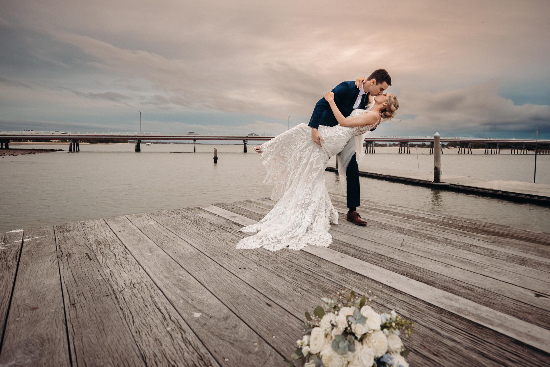 Courtney and Cameron pose on a wooden dock at Sandstone Point Hotel, with Cameron dipping Courtney in her wedding dress as they kiss, a bouquet placed on the dock in the foreground and a bridge visible over the water in the background.