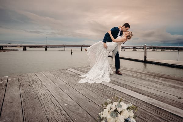 Courtney and Cameron pose on a wooden dock at Sandstone Point Hotel, with Cameron dipping Courtney in her wedding dress as they kiss, a bouquet placed on the dock in the foreground and a bridge visible over the water in the background.