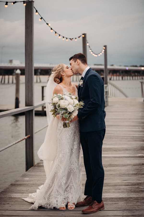 Bride Courtney and groom Cameron kiss on a wooden pier at Sandstone Point Hotel, with string lights overhead and water in the background. Courtney holds a bouquet of white flowers and wears a lace wedding dress with a veil, while Cameron wears a dark suit with a boutonniere.