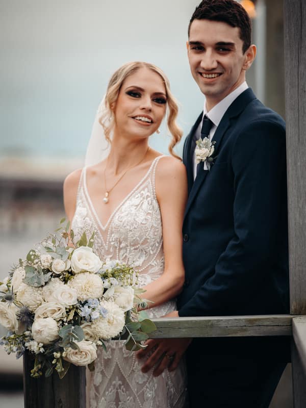 Courtney the bride and Cameron the groom pose together holding a bouquet of white flowers at Sandstone Point Hotel during their couple portraits session.
