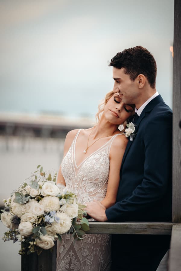Courtney the bride in a lace wedding dress holding a bouquet of white flowers leans her head on Cameron the groom's shoulder as he stands beside her in a dark suit at Sandstone Point Hotel.