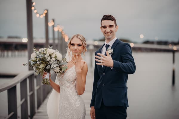 Courtney and Cameron stand on a pier at Sandstone Point Hotel, both showing their wedding rings. Courtney holds a bouquet of white and green flowers while wearing a lace wedding dress, and Cameron is dressed in a dark suit with a boutonniere.