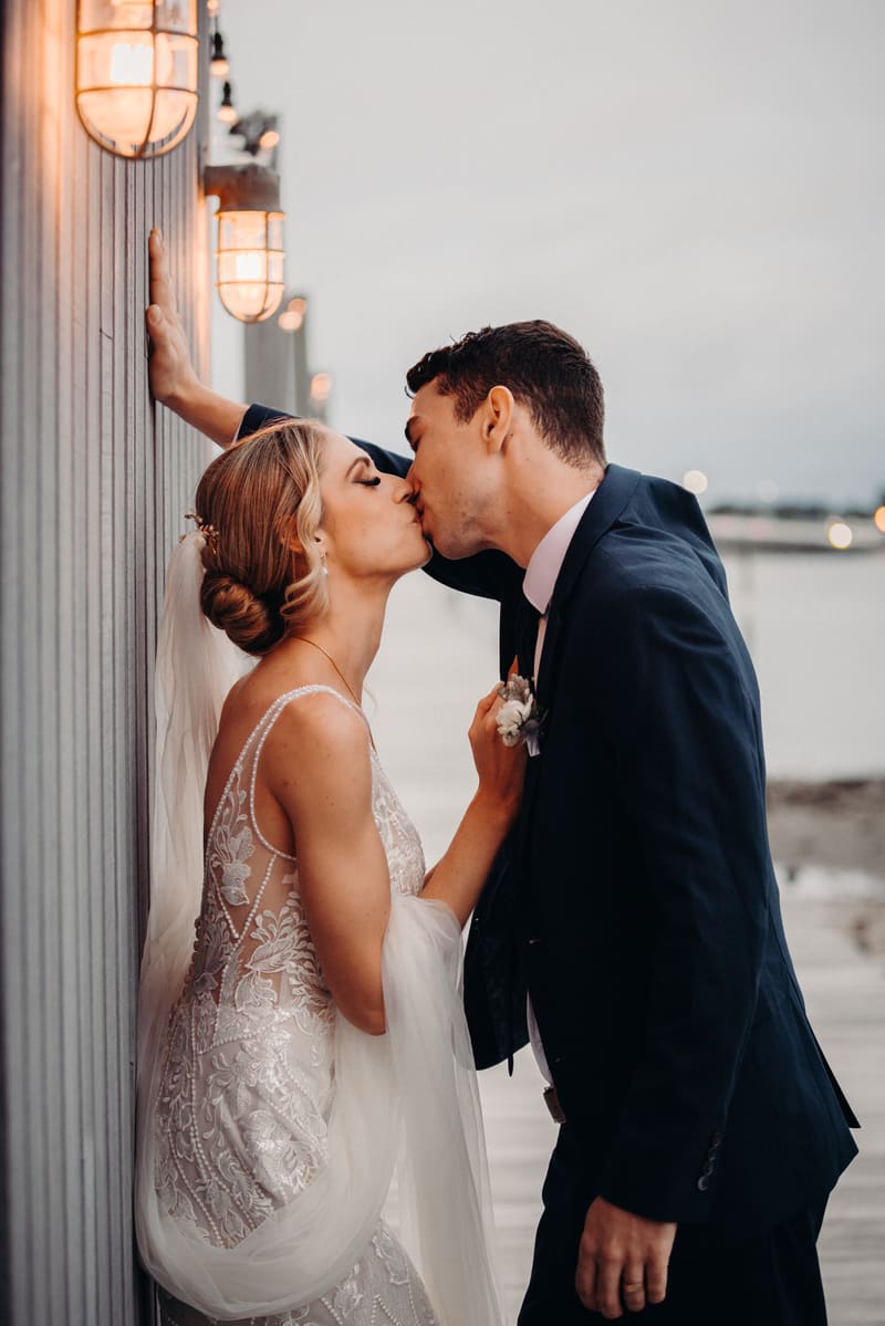 Courtney the bride and Cameron the groom share a kiss during their couple portraits session, standing beside a wall with lit sconces. Wedding photography by She Said Yes.