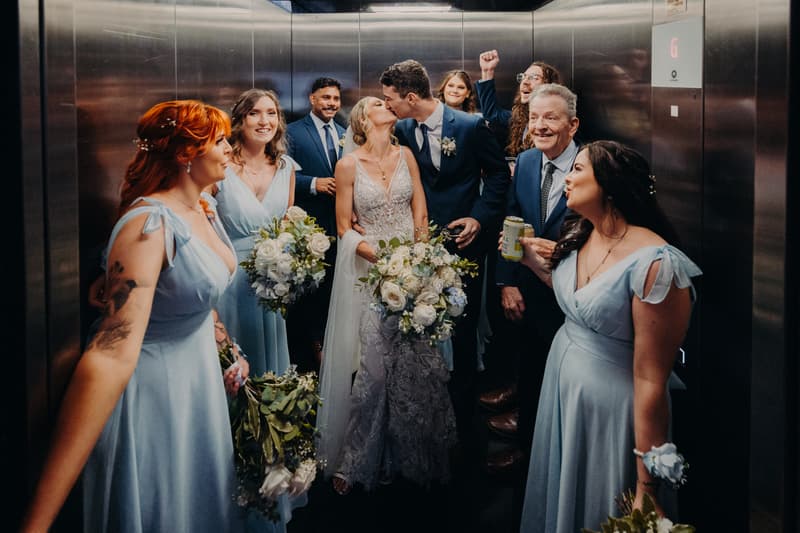 Courtney the bride and Cameron the groom kiss in an elevator at Sandstone Point Hotel surrounded by bridesmaids in light blue dresses holding bouquets, a man in a suit, and an older man holding a can.